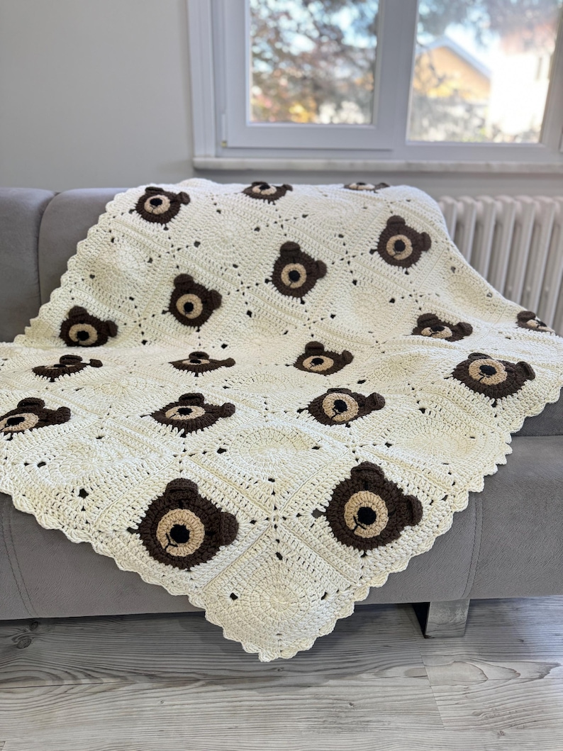 May include: A cream-colored crochet blanket featuring a repeating pattern of square motifs, each decorated with a dark brown bear face. The bear faces consist of a dark brown circular outline with a lighter tan center, small ears, and a black stitched nose.