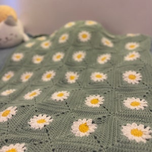 May include: A handmade crochet blanket featuring a daisy pattern. The blanket is primarily sage green with white and yellow daisy appliqués. The square pattern is visible, and the blanket appears soft and cozy.
