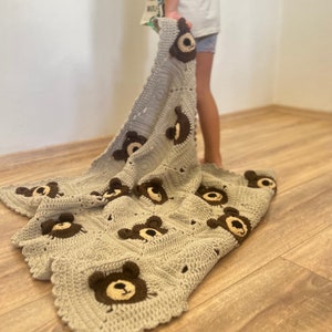 May include: A light gray crochet blanket featuring a repeating pattern of square motifs, each decorated with a dark brown and cream-colored bear face appliqu&eacute;. The blanket has a scalloped edge and is constructed from textured yarn stitches.