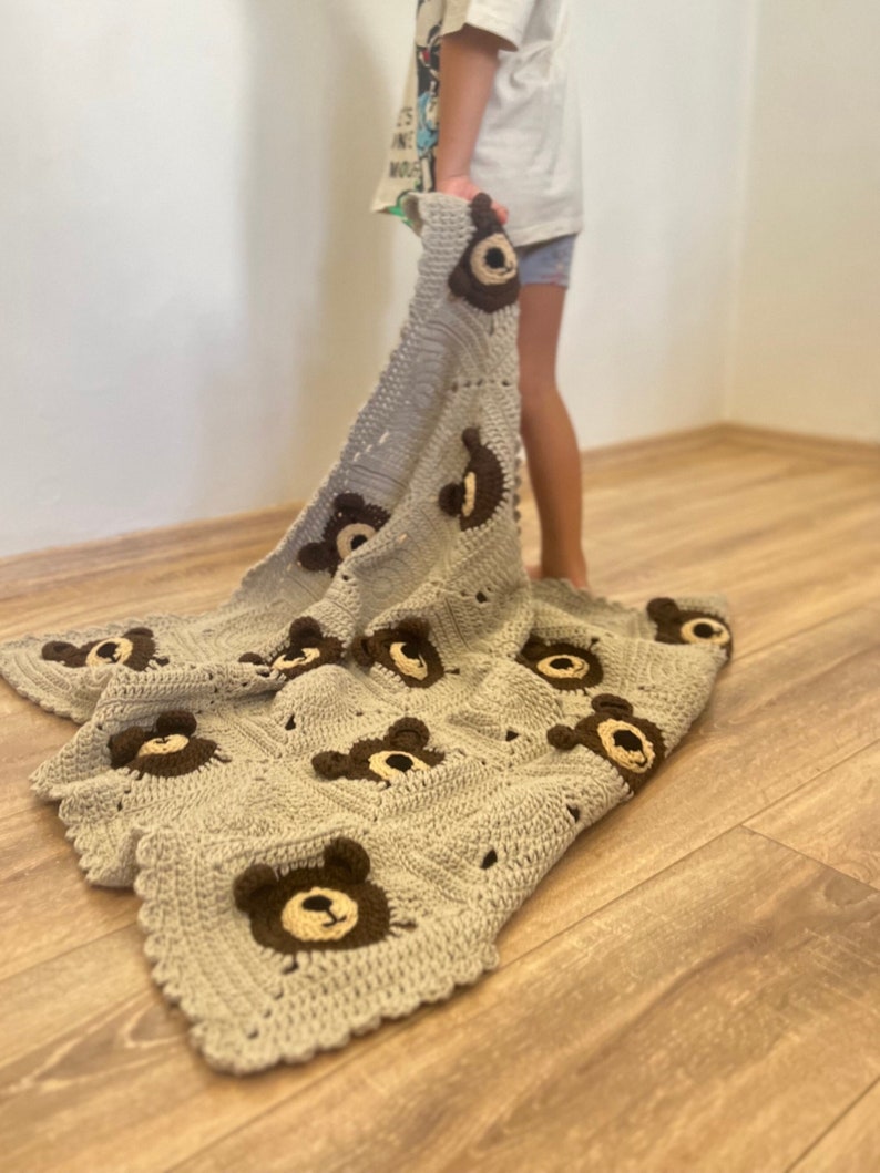 May include: A light gray crochet blanket featuring a repeating pattern of square motifs, each decorated with a dark brown and cream-colored bear face appliqu&eacute;. The blanket has a scalloped edge and is constructed from textured yarn stitches.