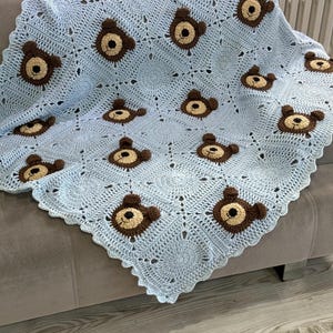 May include: A light blue crochet blanket featuring a repeating pattern of square motifs. Each square contains a central brown and tan bear face with small ears, dark eyes, and a stitched nose. The blanket has a scalloped edge border.