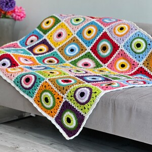 Puede incluir: Una manta de ganchillo colorida con un patrón repetitivo de círculos multicolores con centros negros. La manta está doblada y drapeada sobre un sofá gris claro.