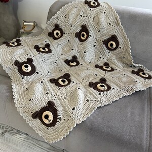 May include: A handmade crochet blanket featuring a repeating pattern of light beige squares and dark brown bear faces. The blanket has a scalloped edge and is constructed from individual squares joined together, showcasing a textured yarn design.