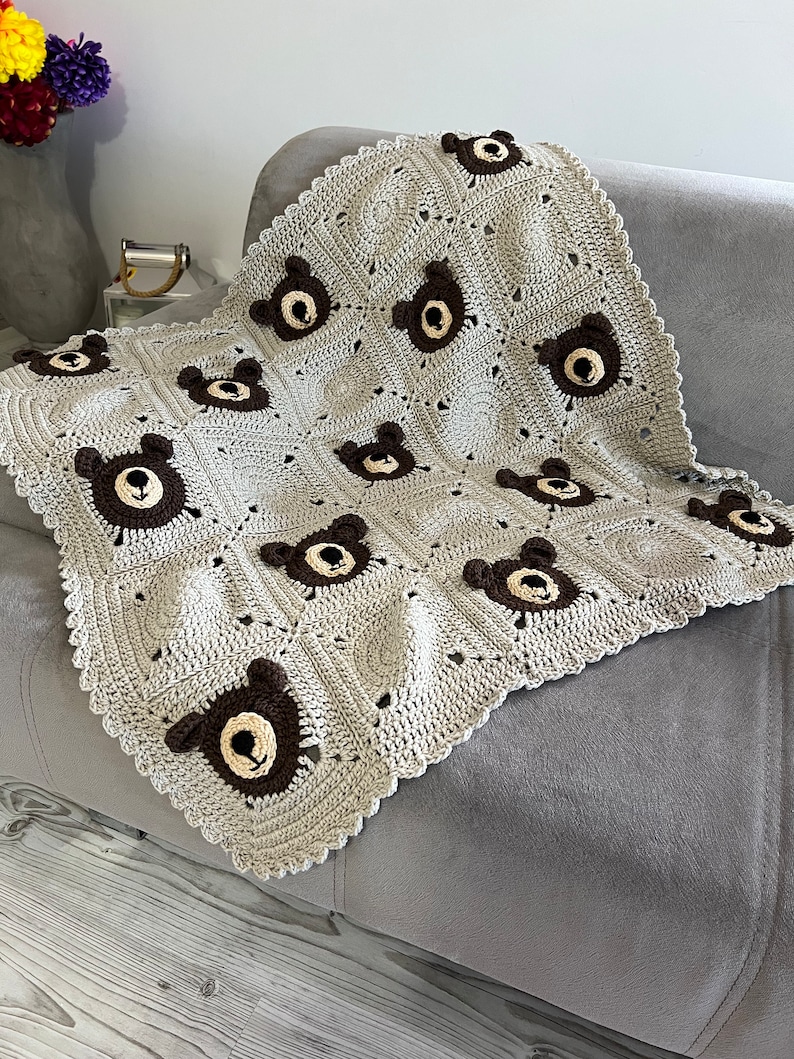 May include: A handmade crochet blanket featuring a repeating pattern of light beige squares and dark brown bear faces. The blanket has a scalloped edge and is constructed from individual squares joined together, showcasing a textured yarn design.