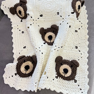 May include: A white crocheted baby blanket with brown bear appliques. The blanket has a scalloped edge and is made with a simple stitch pattern.