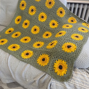 May include: Handmade crochet blanket featuring a repeating sunflower pattern. The blanket has a green background with yellow and brown sunflowers. The blanket is draped over a gray patterned couch.
