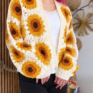 May include: Cream-colored crochet cardigan featuring large sunflower motifs in shades of yellow and brown. The open-front sweater has long sleeves and a relaxed fit. The sunflower pattern is repeated across the front and sleeves.