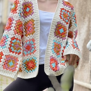 May include: A white crochet cardigan with a granny square pattern in shades of pink, orange, and blue. The cardigan has a V-neck and long sleeves.