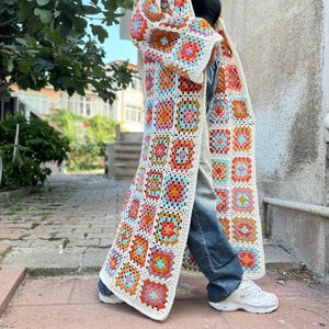 May include: A long, white crochet cardigan with a colorful geometric pattern. The cardigan has a loose fit and long sleeves. The model is wearing the cardigan over a black top and blue jeans.