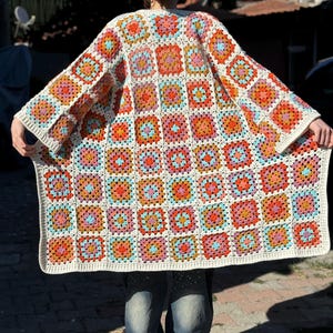 May include: A white crochet cardigan with a granny square pattern in shades of orange, pink, blue, and brown. The cardigan has a long, flowing silhouette and is perfect for layering.