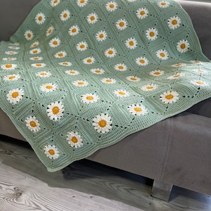 Puede incluir: Una manta de crochet con fondo verde y margaritas blancas con centros amarillos. La manta está drapeada sobre un sofá gris.