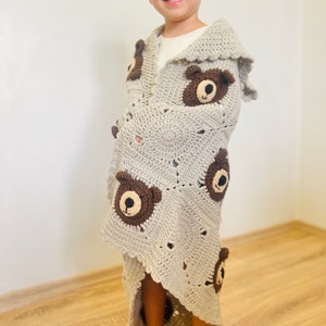 May include: A grey crocheted hooded blanket with brown bear faces. The blanket is draped over a child.