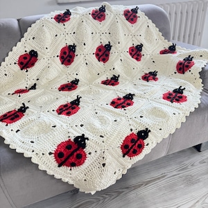 Puede incluir: Una manta de ganchillo blanca con un diseño de mariquita. La manta está hecha con hilo blanco y presenta mariquitas rojas y negras.