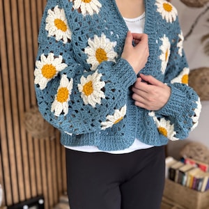 May include: A blue crochet cardigan featuring large daisy flower appliques with white petals and yellow centers. The open-front sweater has long sleeves and a scalloped hem. The garment is handmade and has a relaxed fit.