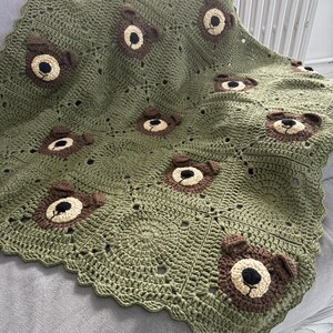 May include: A sage green crochet blanket featuring a repeating pattern of square motifs. Several squares are embellished with three-dimensional brown bear faces, each crafted with a cream-colored muzzle, black nose, and small rounded ears.