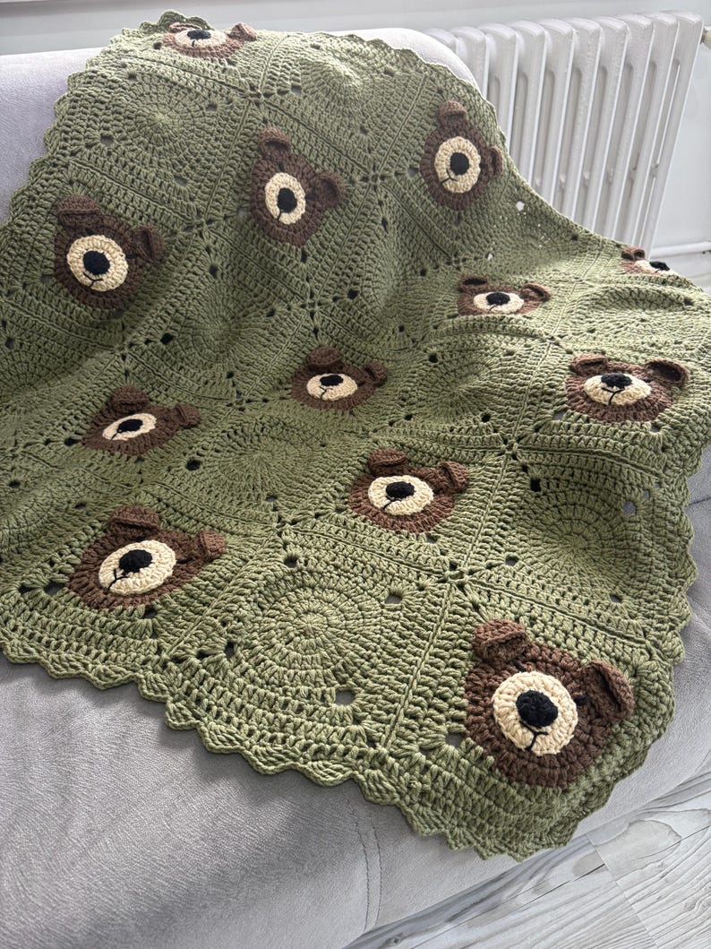 May include: A sage green crochet blanket featuring a repeating pattern of square motifs. Several squares are embellished with three-dimensional brown bear faces, each crafted with a cream-colored muzzle, black nose, and small rounded ears.