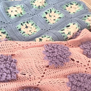 Könnte beinhalten: Zwei handgefertigte Häkeldecken. Die obere Decke ist hellblau mit weißen und mintgrünen Blumenquadraten. Die untere Decke ist rosa mit großen, strukturierten Lavendel-Blütenakzenten. Die Decken sind weich und gemütlich.
