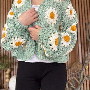 Puede incluir: Un cárdigan de crochet verde claro con grandes aplicaciones de flores de margarita. Las flores tienen pétalos blancos y centros amarillos. Las mangas son largas y el cárdigan está abierto por delante.