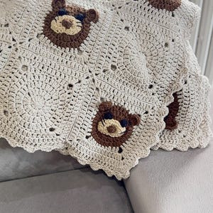 May include: A handmade crochet blanket featuring a repeating pattern of bear faces. The blanket is primarily beige with brown bear details. The edges have a scalloped trim. This cozy blanket is perfect for a nursery or as a decorative throw.
