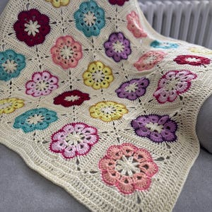 May include: A cream-coloured, crocheted blanket with colourful floral designs in red, pink, yellow, teal, and purple. The blanket is draped over a grey surface, showcasing the intricate stitchwork and vibrant colour palette.