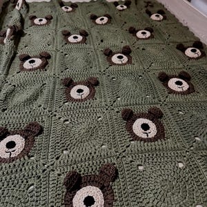 May include: A handmade crochet blanket featuring a repeating pattern of olive green squares, each centered with a brown and beige bear face motif. The bear faces include dark brown ears, a light beige muzzle, and a black stitched nose and mouth.