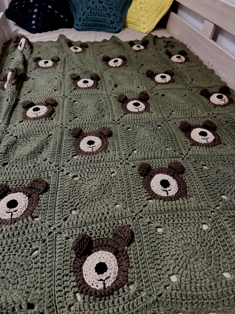 May include: A handmade crochet blanket featuring a repeating pattern of olive green squares, each centered with a brown and beige bear face motif. The bear faces include dark brown ears, a light beige muzzle, and a black stitched nose and mouth.