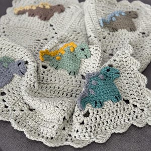 Może przedstawiać: Jasnoszary, szydełkowy koc z falistym brzegiem. Koc posiada cztery kwadraty, każdy z inną kolorową aplikacją dinozaura: brązowym, niebieskim, zielonym i turkusowym. Dinozaury mają żółte lub niebieskie akcenty.