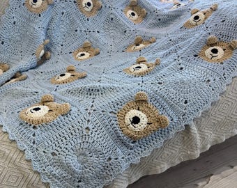 May include: A light blue crocheted blanket featuring a repeating pattern of teddy bear faces. The bears have tan ears and faces with black and white eyes. The blanket is draped over a sofa, showcasing its handmade design.