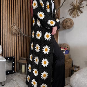 May include: A long, black crochet cardigan featuring a daisy pattern. The daisies have white petals and yellow centers. The cardigan is knee-length and has long sleeves. The garment is worn over a white top and black pants.