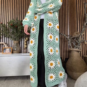 May include: A long, open-front crochet cardigan in a light green color, adorned with white and yellow daisy flower motifs. The cardigan has long sleeves and a knee-length design. The garment is made of a soft, open-weave material.