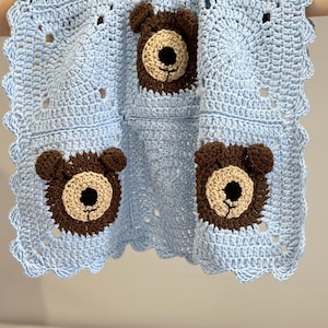 May include: A light blue crochet blanket featuring three square panels with dark brown bear faces. Each bear has a beige muzzle, black nose, and small rounded ears. The blanket is finished with a scalloped edge border in light blue yarn.
