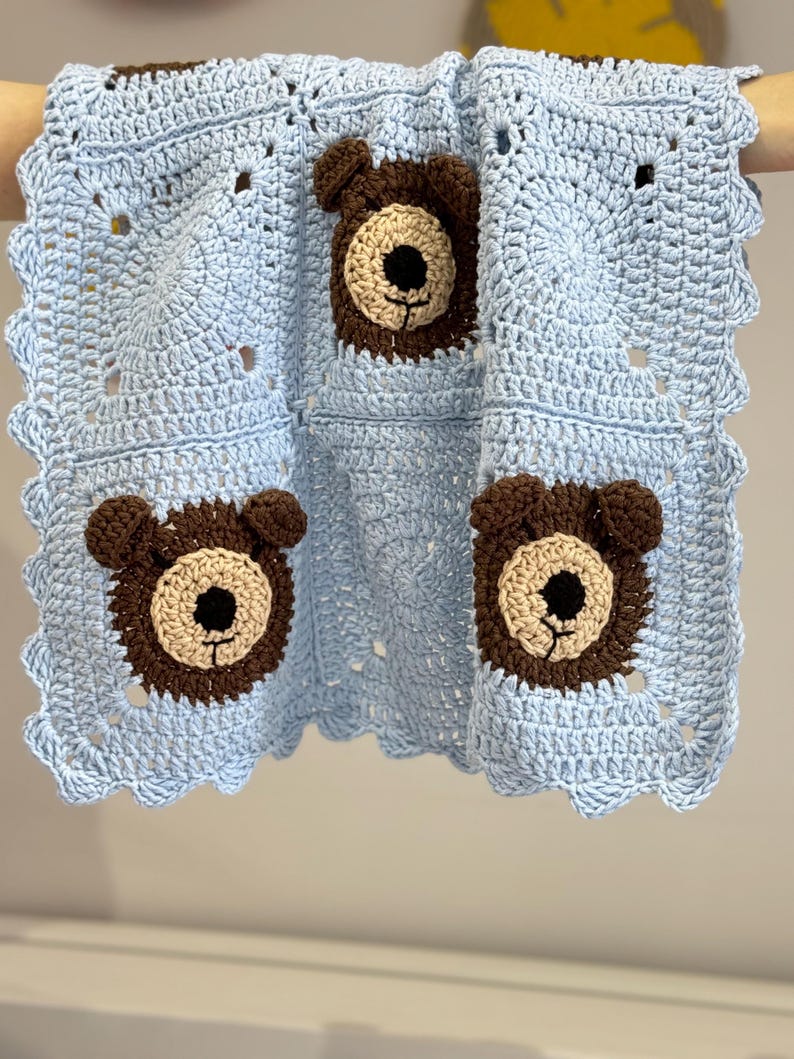 May include: A light blue crochet blanket featuring three square panels with dark brown bear faces. Each bear has a beige muzzle, black nose, and small rounded ears. The blanket is finished with a scalloped edge border in light blue yarn.