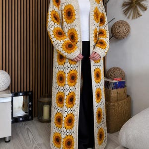 May include: A long, open-front crochet cardigan featuring a sunflower pattern. The cardigan is cream-colored with yellow and brown sunflower motifs. The garment is knee-length and has a loose, flowing fit, perfect for layering.