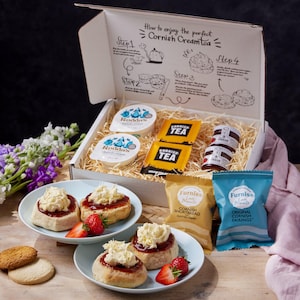 May include: A white box filled with a Cornish cream tea gift set. The box contains two tubs of clotted cream, two tea bags, a jar of jam, a packet of shortbread, and a packet of Cornish fairings. The box is open and the contents are visible. There are also two plates with scones, clotted cream, jam, and strawberries.