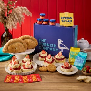 May include: A blue shipping box branded The Cornish Company with a seagull logo, surrounded by scones topped with clotted cream and strawberry jam, two pasties, small jam jars, tea bags, and a yellow sign reading cream on top and dare to dunk.