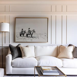 May include: A white couch with three pillows, two dark gray and one light beige, sits in front of a white wall with a framed black and white drawing of two people riding horses. A gold floor lamp with a white lampshade is in the corner of the room.