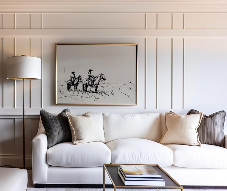 May include: A white couch with three pillows, two dark gray and one light beige, sits in front of a white wall with a framed black and white drawing of two people riding horses. A gold floor lamp with a white lampshade is in the corner of the room.