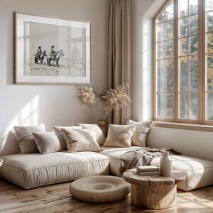 May include: A light beige sectional sofa with plush cushions and a wooden coffee table. The sofa is positioned in front of a large window with a view of trees. A black and white framed print of two people riding horses hangs on the wall.