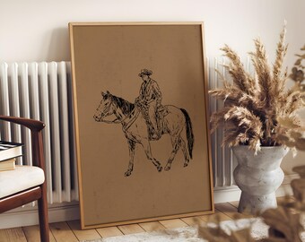 Może przedstawiać: Oprawiony wydruk z czarno-białą ilustracją osoby na koniu. Grafika jest umieszczona na beżowym tle i oprawiona w jasne drewno. Wydruk opiera się o ścianę w pobliżu krzesła i wazonu z suszonymi roślinami.