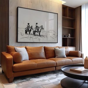 May include: A brown leather sofa with three cushions and a black coffee table in front of it. A black framed picture of two cowboys riding horses hangs on the wall behind the sofa.