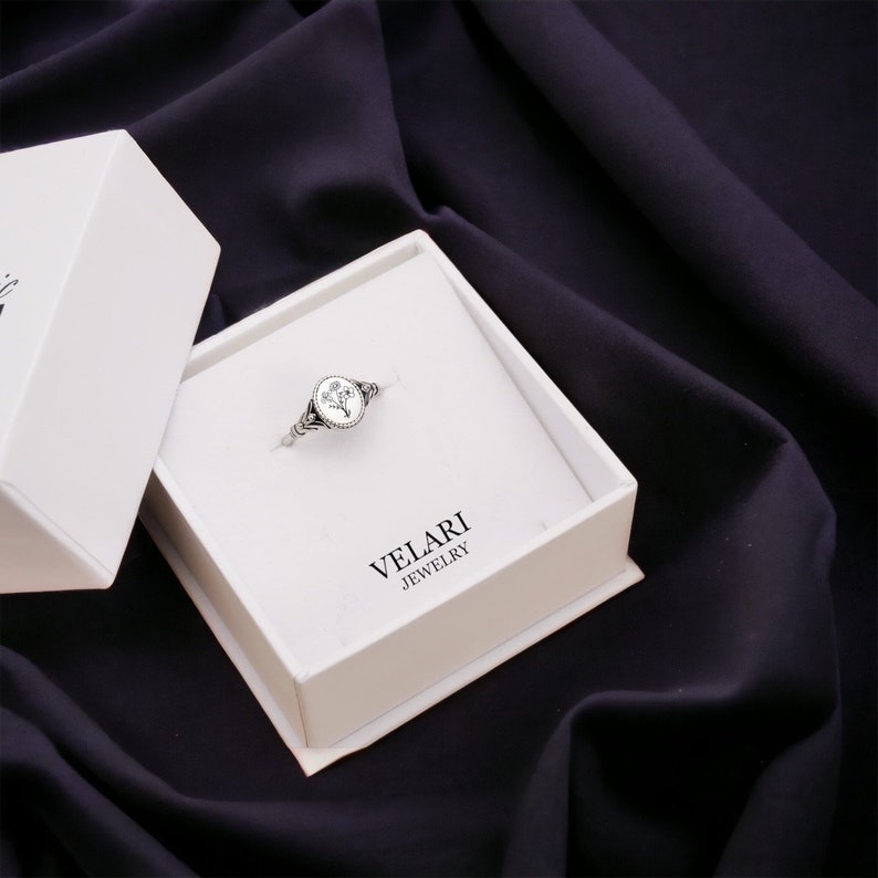 May include: A silver ring with a white oval stone featuring a floral design. The ring is displayed in a white box with the text "VELARI JEWELRY".