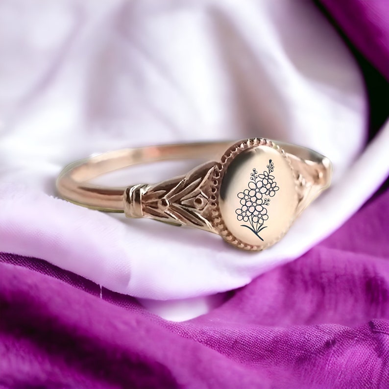 May include: A gold signet ring with an oval shaped bezel featuring an engraved floral design. The ring is set on a white and purple fabric background.