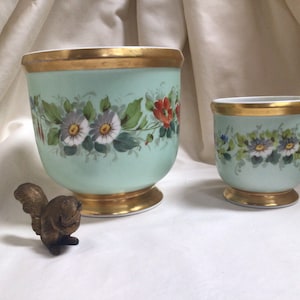 May include: Two matching, pale green porcelain planters with gold rims and bases. Each planter features a floral design in red, white, and blue. A small bronze squirrel figurine is positioned in front of the planters.