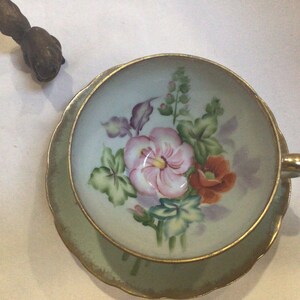 Op de afbeelding: Een vintage kop en schotel set met een bloemenontwerp. De kop en schotel zijn lichtgroen met gouden rand. De kop heeft een roze en oranje bloemenboeket. Een decoratief houten object bevindt zich in de linkerbovenhoek.