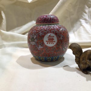 May include: A red and gold porcelain jar with intricate floral designs and a white circle with the Chinese character "金" (jin) in the center. The jar has a lid and is sitting on a white fabric surface.