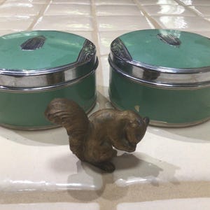 以下が含まれることがあります： シルバーの縁と蓋が付いた、2つのヴィンテージのティール色のパウダージャー。小さなブロンズ色のリスの置物がジャーの前に配置されています。ジャーは丸い形で、蓋には装飾的なシルバーのアクセントが付いています。