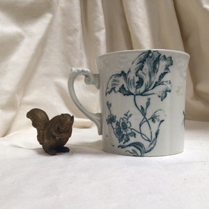 May include: White ceramic mug with a blue floral pattern and a decorative handle. A small bronze squirrel figurine is beside the mug. The mug features a large flower and smaller floral accents.