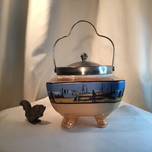 May include: A vintage ceramic cookie jar with a silver lid and handle. The jar is decorated with a blue and white scene of a beach with boats and people. The jar has three feet and is in excellent condition.