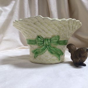 May include: A pale yellow ceramic basket planter with a green bow. A small, brown squirrel figurine is positioned next to the planter. The planter has a woven texture and a scalloped edge, suitable for home decor.
