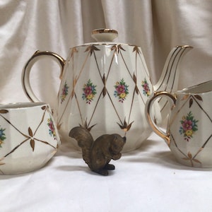 以下が含まれることがあります： ティーポット、クリーマー、砂糖入れのあるヴィンテージティーセット。白い磁器には金色のアクセントと花の模様があります。小さなブロンズのリスの置物がティーセットの前に置かれています。